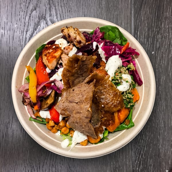 A colorful bowl of Mediterranean food with sliced meat, chicken, vegetables, and sauce on a dark wooden table.