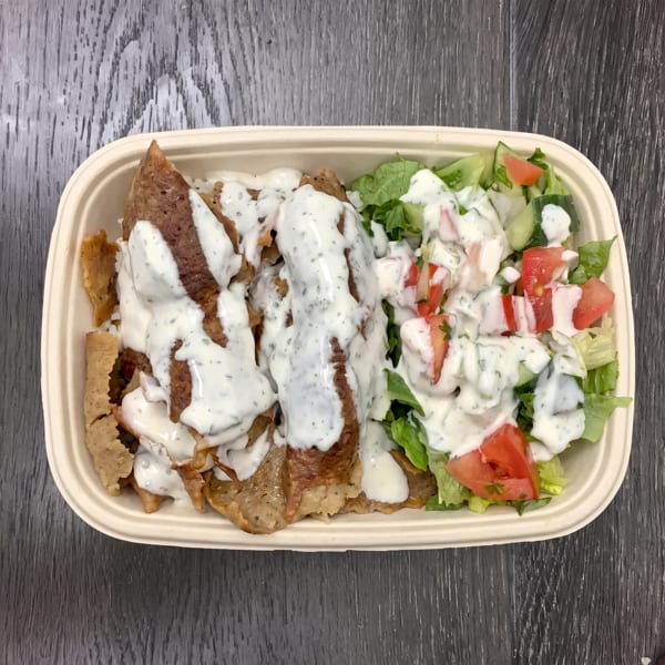 A takeout container with gyro meat, white sauce, and a side salad with tomatoes and cucumbers.