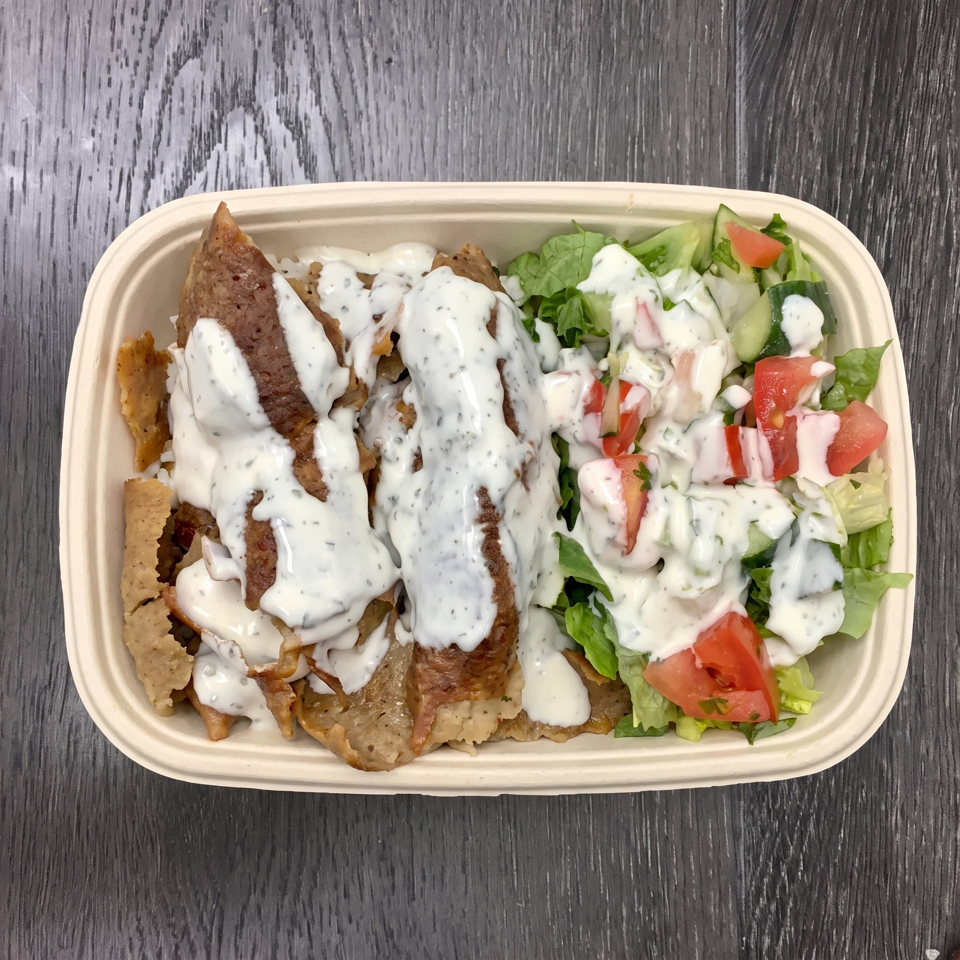 A takeout container with gyro meat, white sauce, and a side salad with tomatoes and cucumbers.