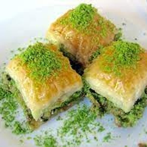 Three golden baklava pastries topped with green pistachios, arranged on a white plate with scattered crumbs.