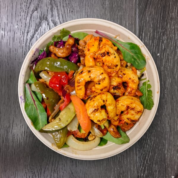A bowl of grilled shrimp, colorful bell peppers, onions, and mixed greens on a dark wooden surface.