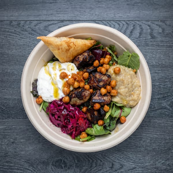 A colorful Mediterranean bowl with grilled meat, chickpeas, yogurt, red cabbage, and a pastry.