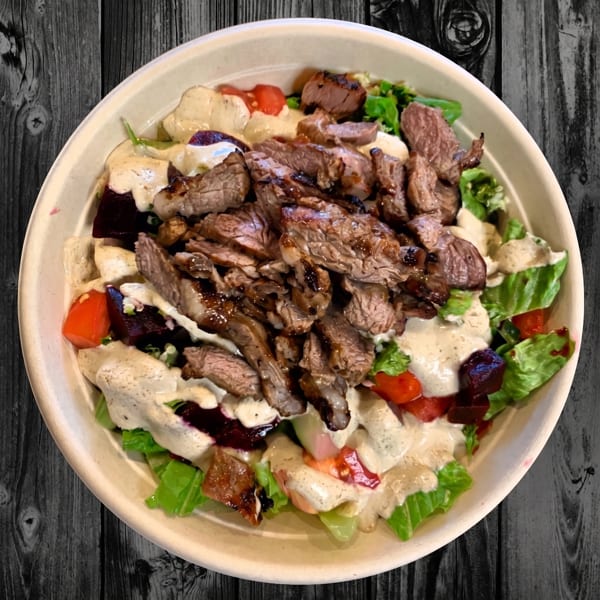 A close-up of a salad bowl filled with grilled steak, lettuce, tomatoes, and a creamy dressing.